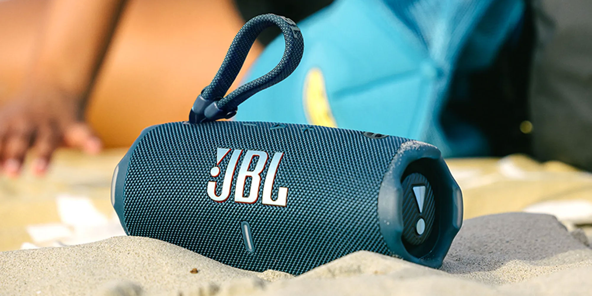 A small JBL speaker is nestled in the sand on a beach, while figures chat in the background