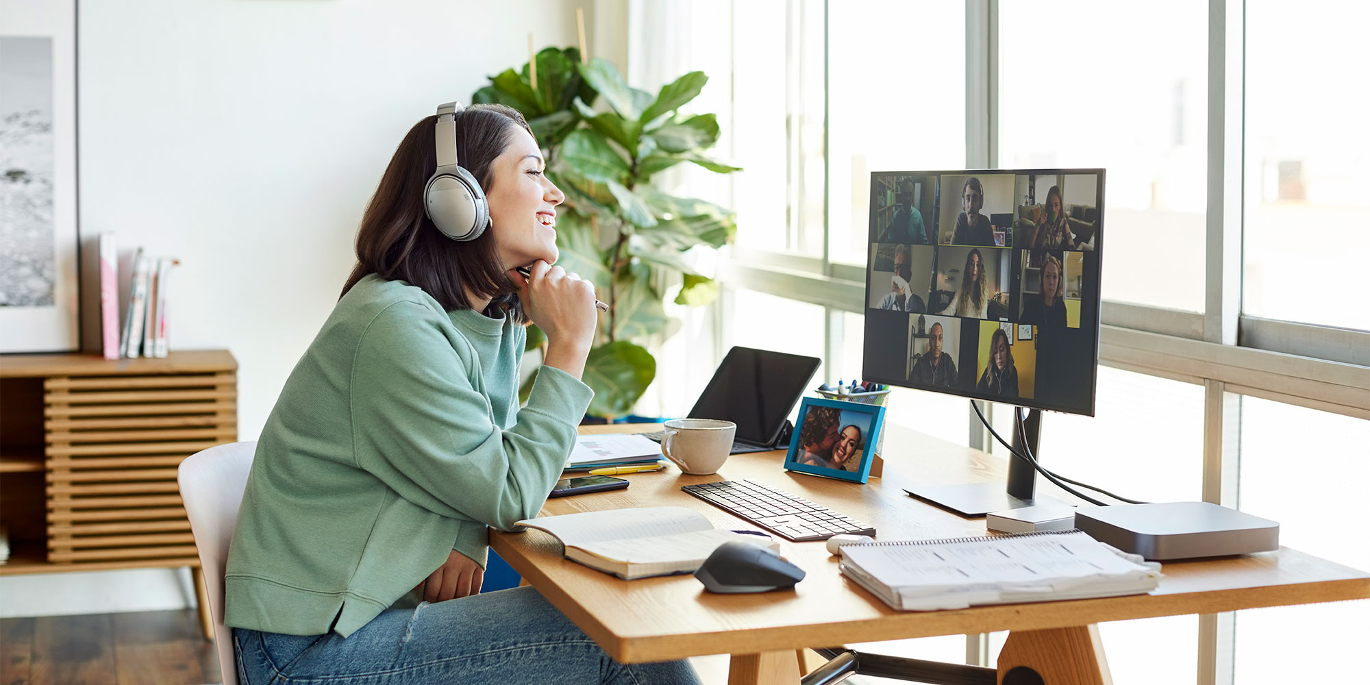 Woman with headphones smiles while participating in a video call on a monitor, surrounded by books and a plant.