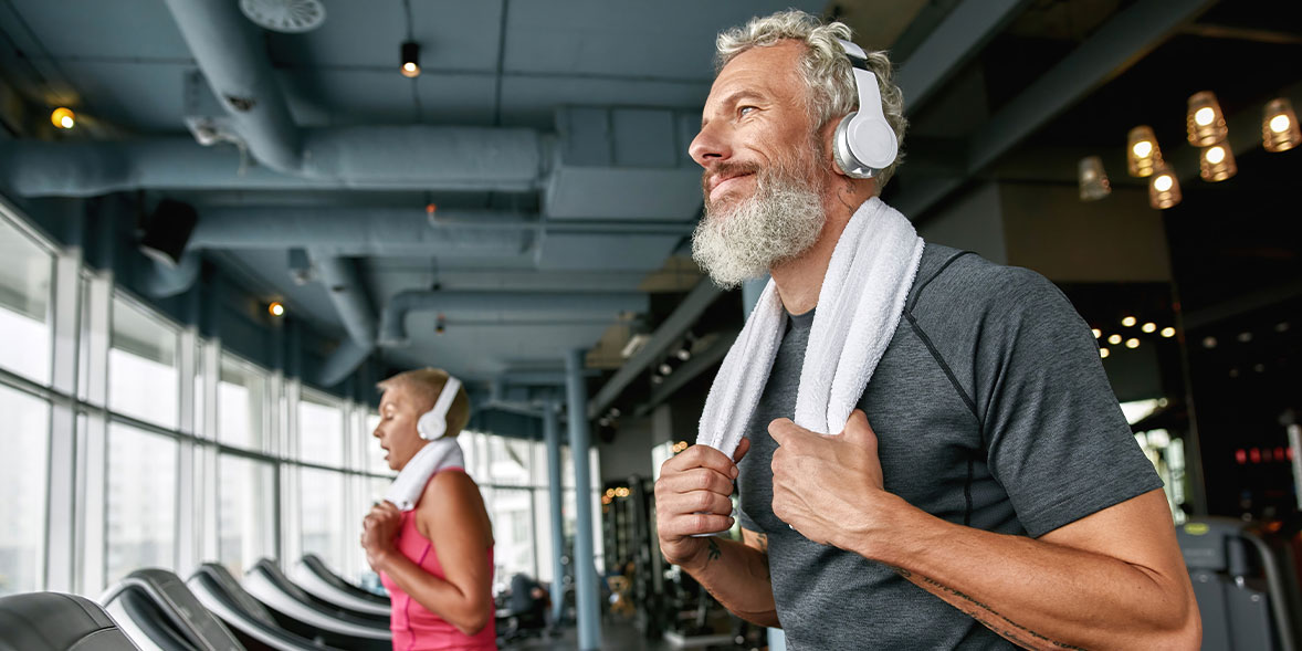Man with a beard and headphones smiles while holding a towel, alongside a woman in workout attire, both in a gym.