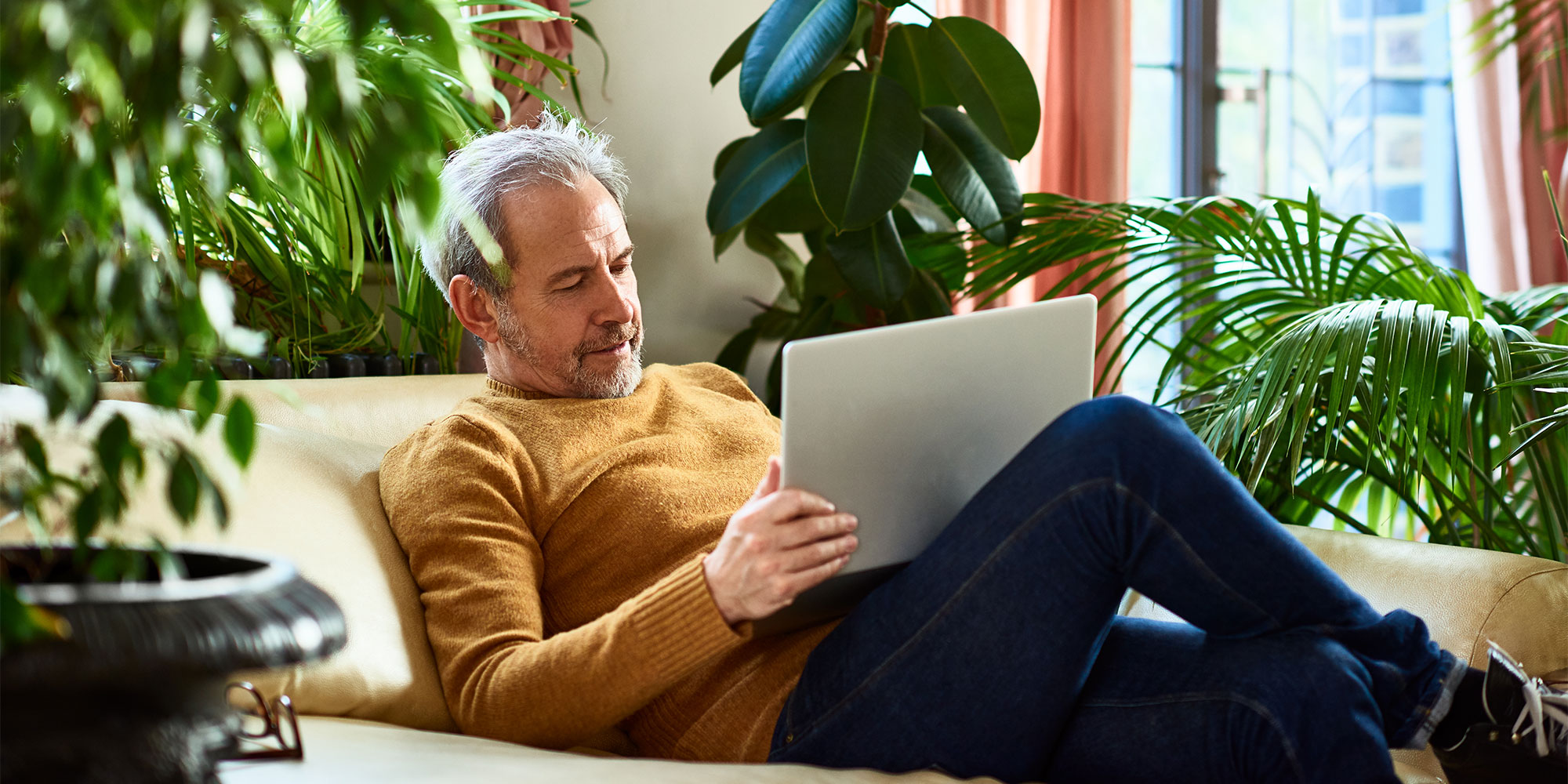 Man sits on a couch, using a laptop, surrounded by lush indoor plants and natural light.