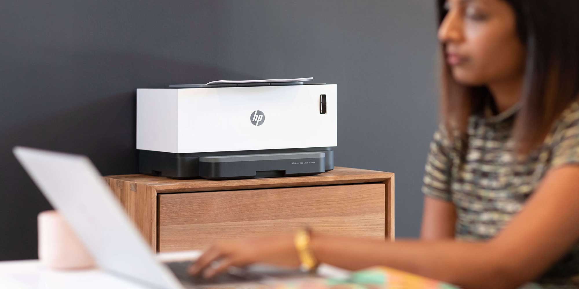 A person holds a colorful printed photo beside an HP printer on a wooden table, with stationery in the background.