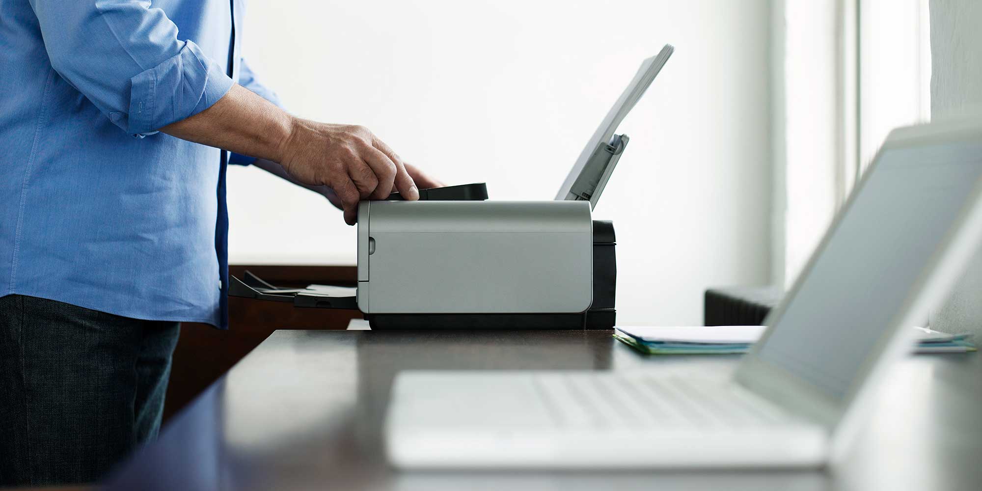 A man in a light blue shirt places a document into a multi-function printer, preparing to scan or copy it.
