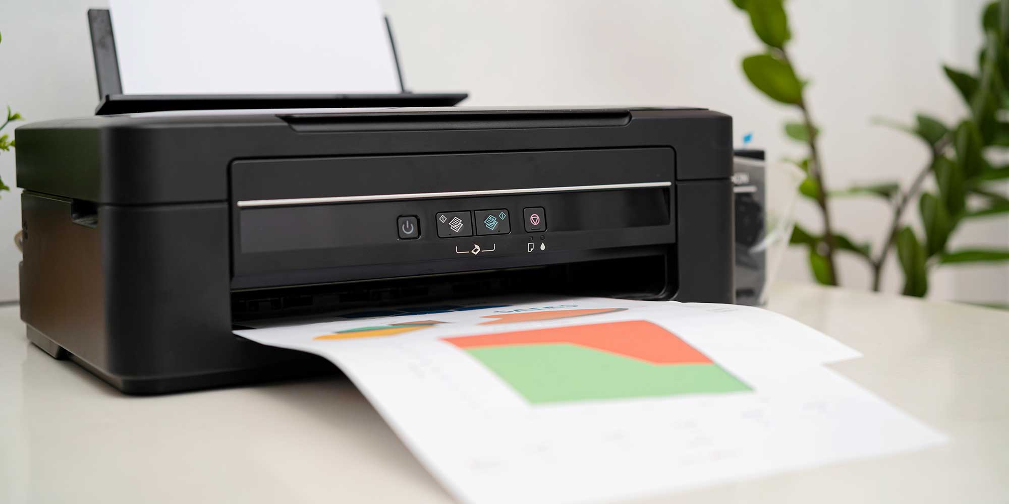 A person in a blue shirt uses a printer, placing a document inside it on a wooden desk with a laptop nearby.