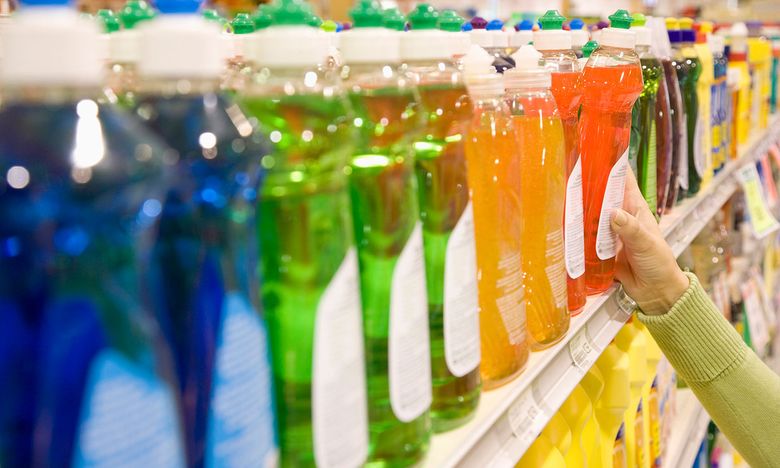 A hand reaches for a bottle of liquid detergent among various colored bottles lined up on a store shelf.