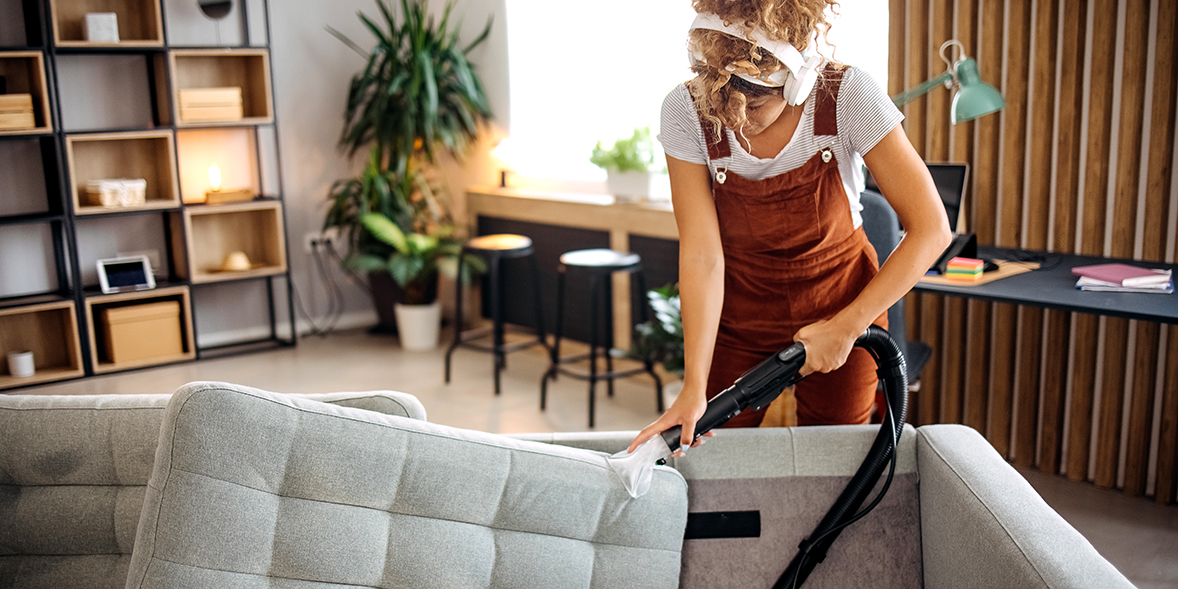 A person vacuum cleans a light grey couch, wearing headphones and a brown jumpsuit, in a bright, modern living room.