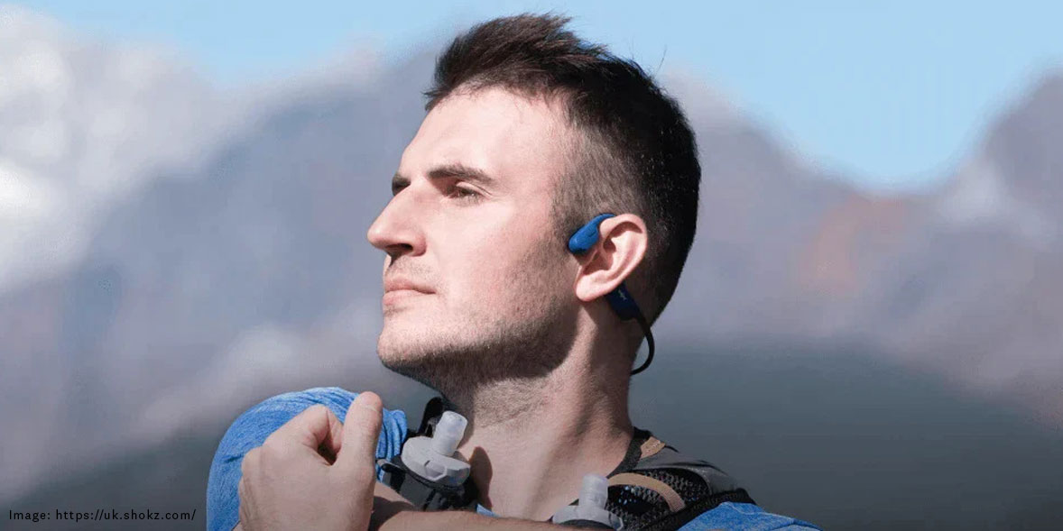 Man with short hair and blue headphones stands outdoors, looking confidently into the distance against a mountain backdrop.
