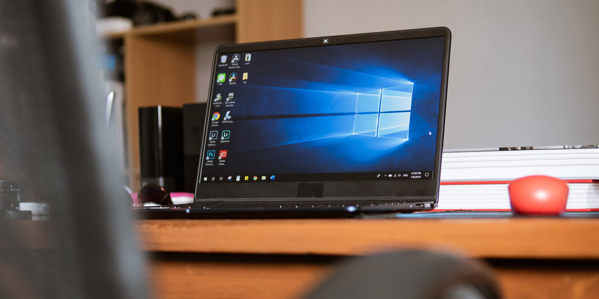 Laptop on a wooden desk displays the Windows interface, with a red object and books in the background.