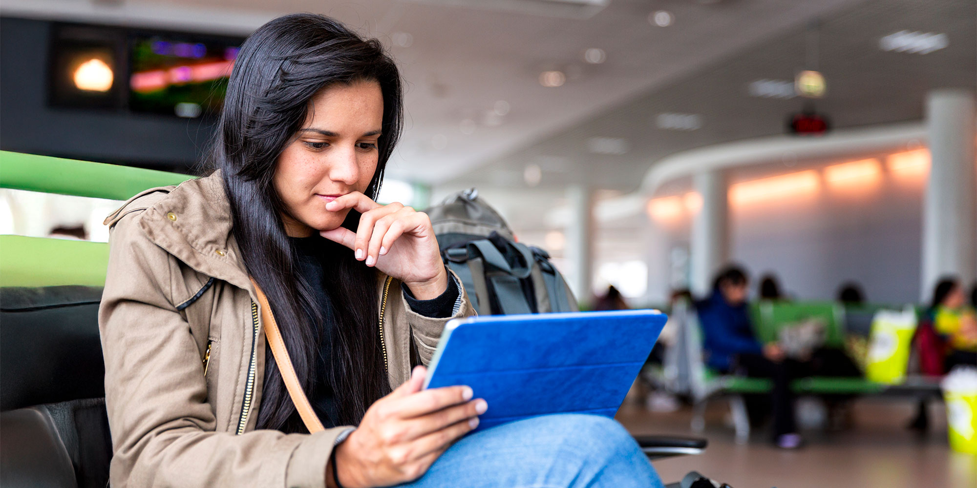 A young woman with long dark hair sits in an airport, focused on a tablet, surrounded by fellow travelers in the background.
