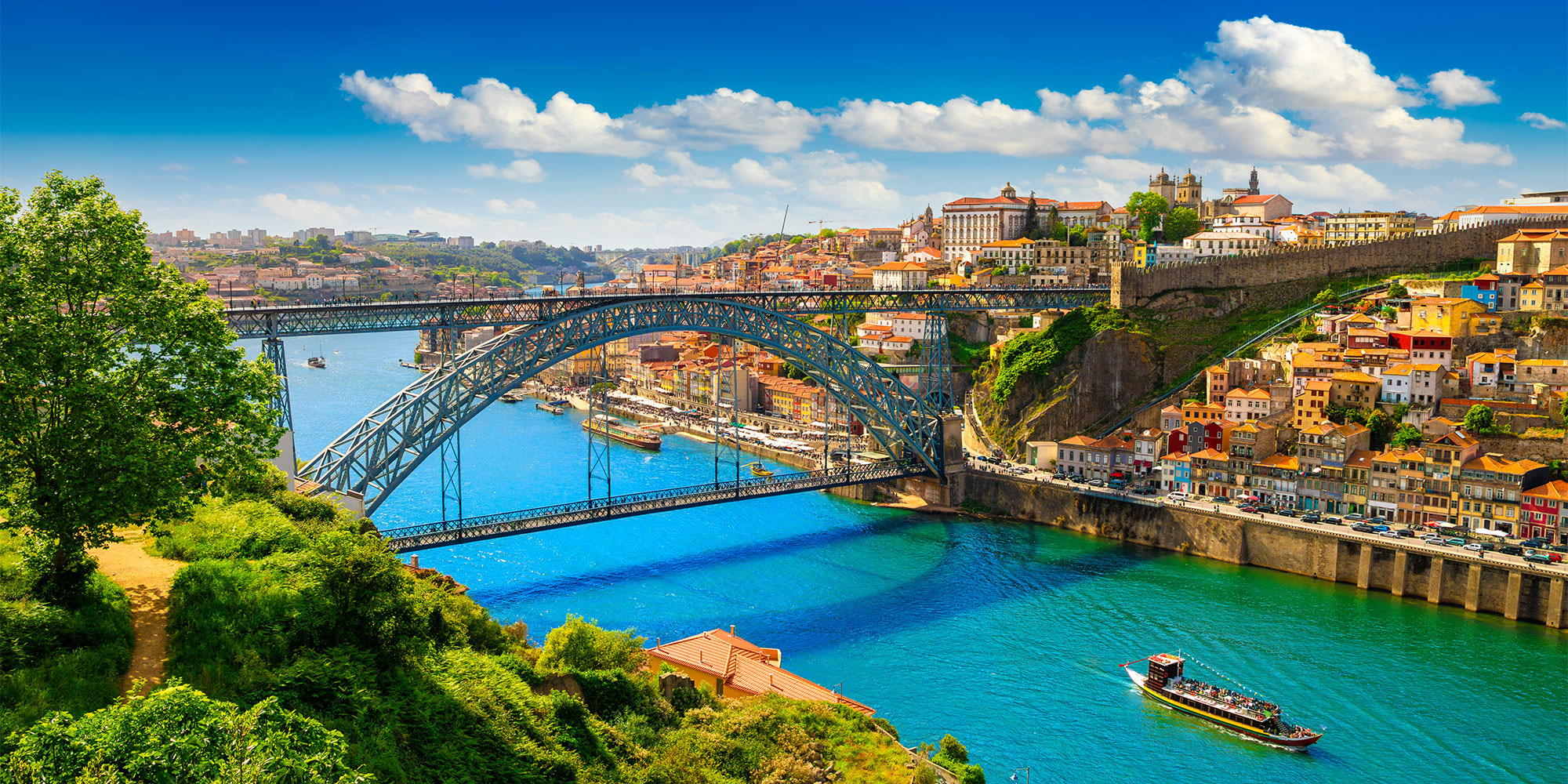 Luxurious blue river under a steel bridge, vibrant houses lining the banks, and a bright sky filled with scattered clouds.