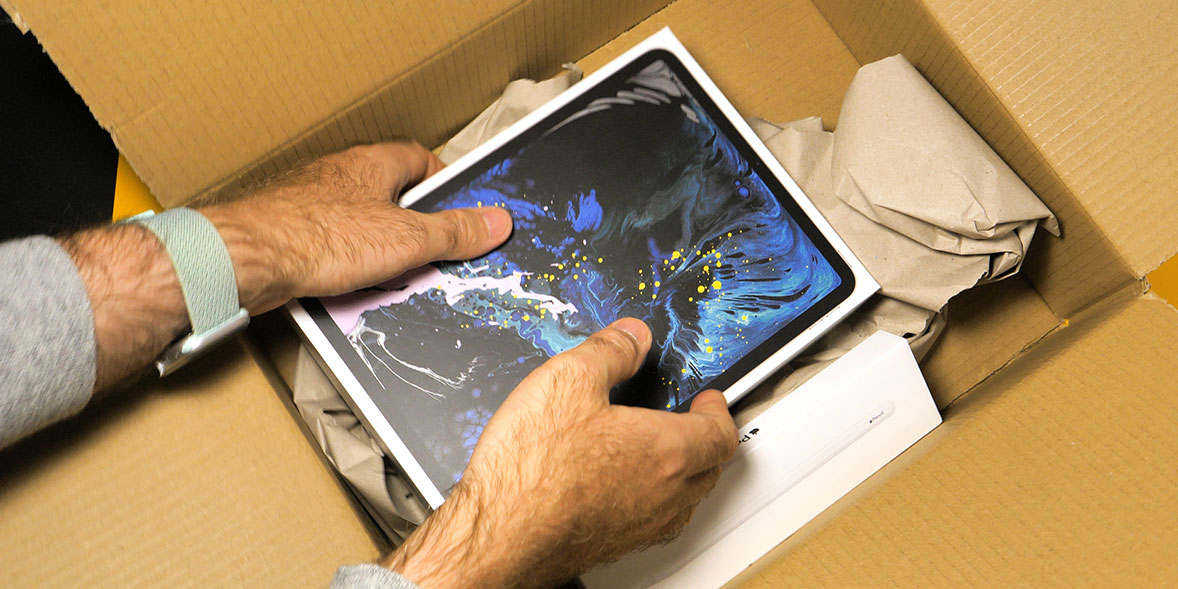 Hands carefully unpack a tablet from a cardboard box filled with crumpled paper, resting next to a white device case.