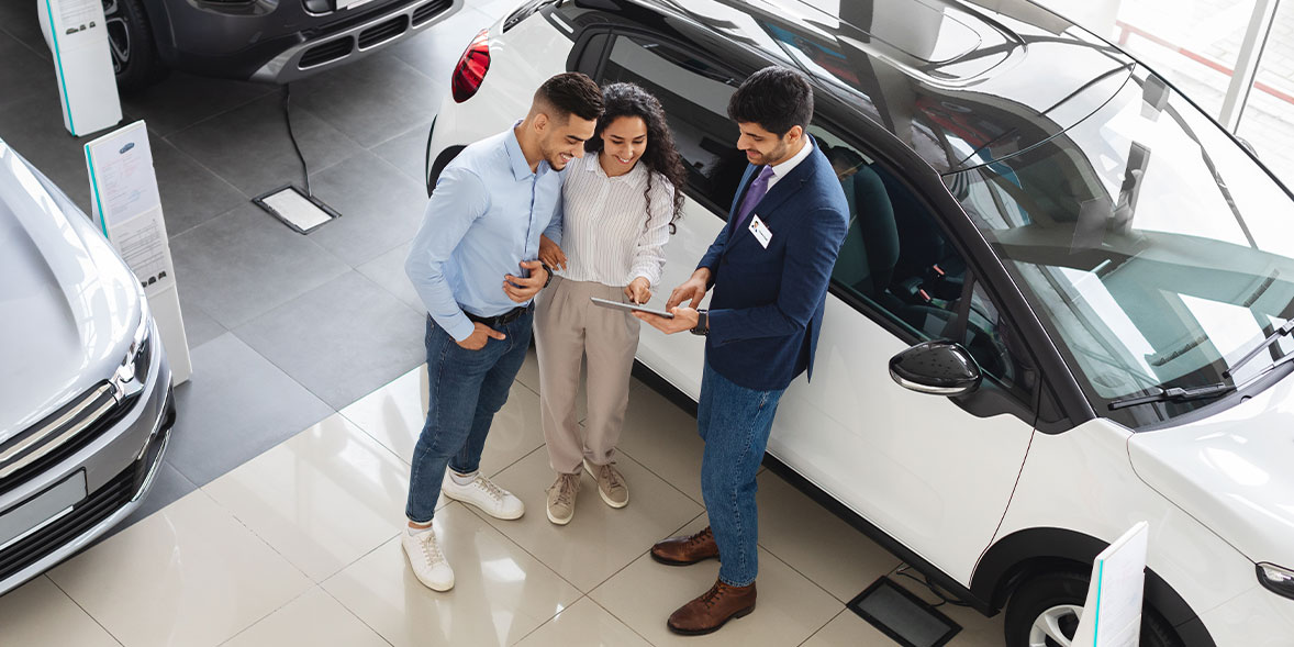 Three people, a salesman and a couple, are discussing details on a tablet inside a car showroom next to a white car.