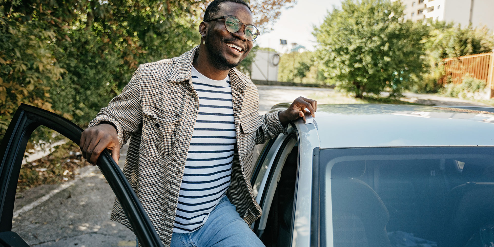 Smiling man in a striped shirt and plaid jacket leans against a car in a sunlit, green outdoor setting.