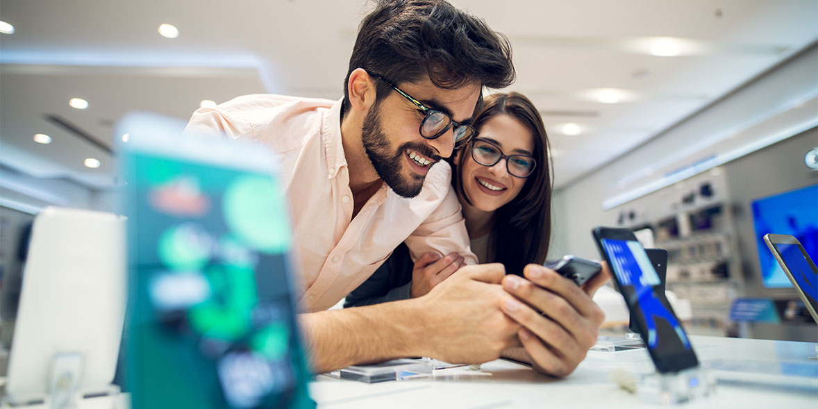 A couple smiles while looking at a smartphone in a tech store, surrounded by various devices on display.