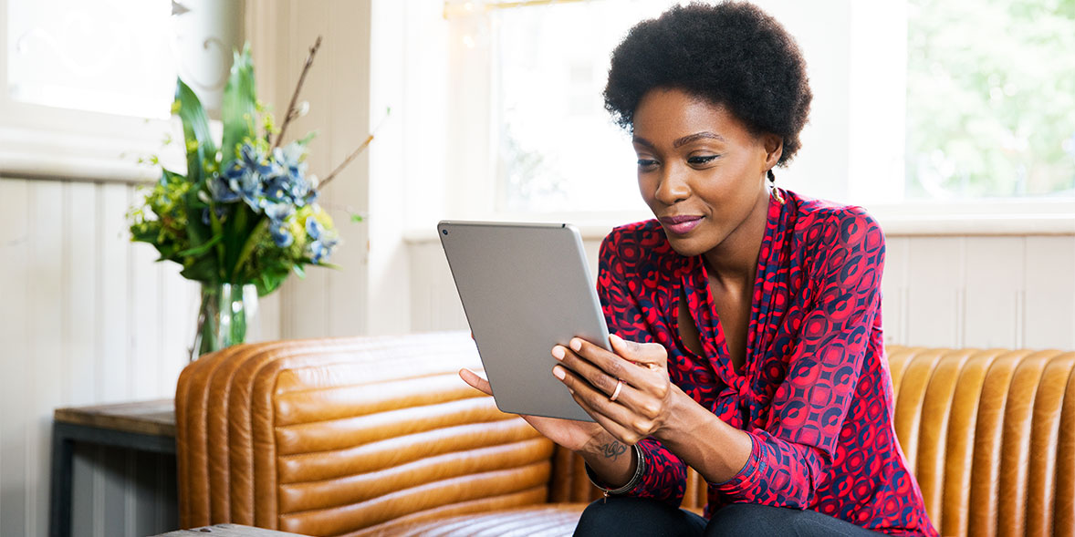 Woman with short, curly hair on a brown leather couch, smiling while holding a tablet. A vase of flowers in the background.