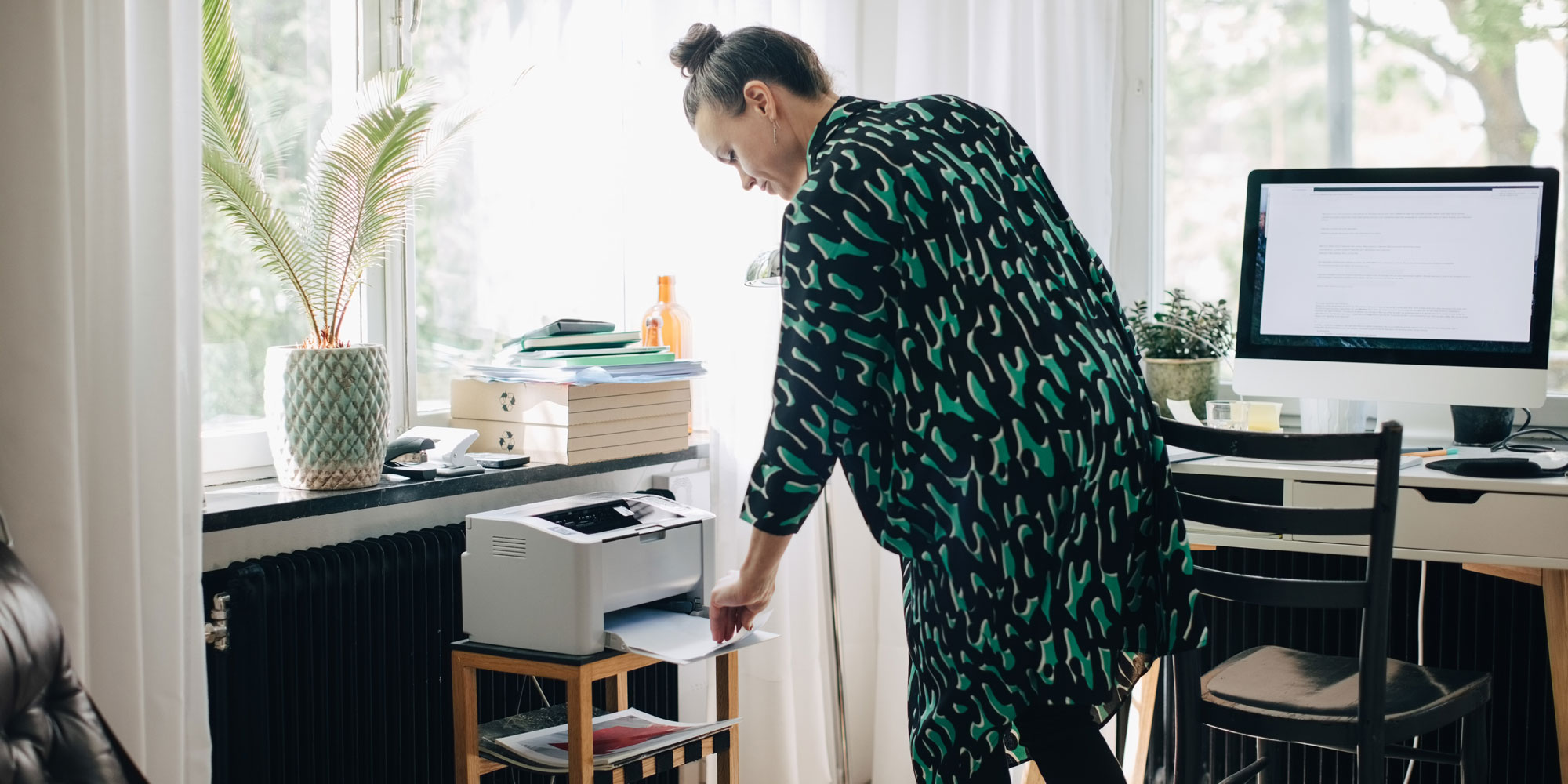 A person in a patterned green and black robe retrieves paper from a printer in a bright home office with a desk and plants.