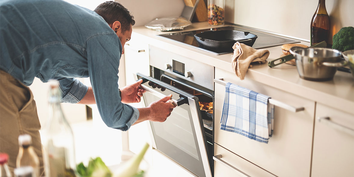 Man in a denim shirt bends down to open an oven, with a kitchen towel hanging nearby and cooking utensils on the counter.