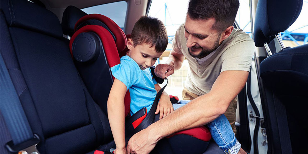 A man helps a young boy secure his seatbelt in a car seat, smiling in a sunny vehicle interior.
