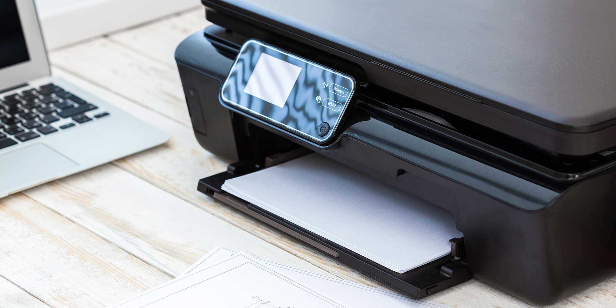 A black printer with a control panel, displaying a blank screen, rests on a wooden surface near a laptop.