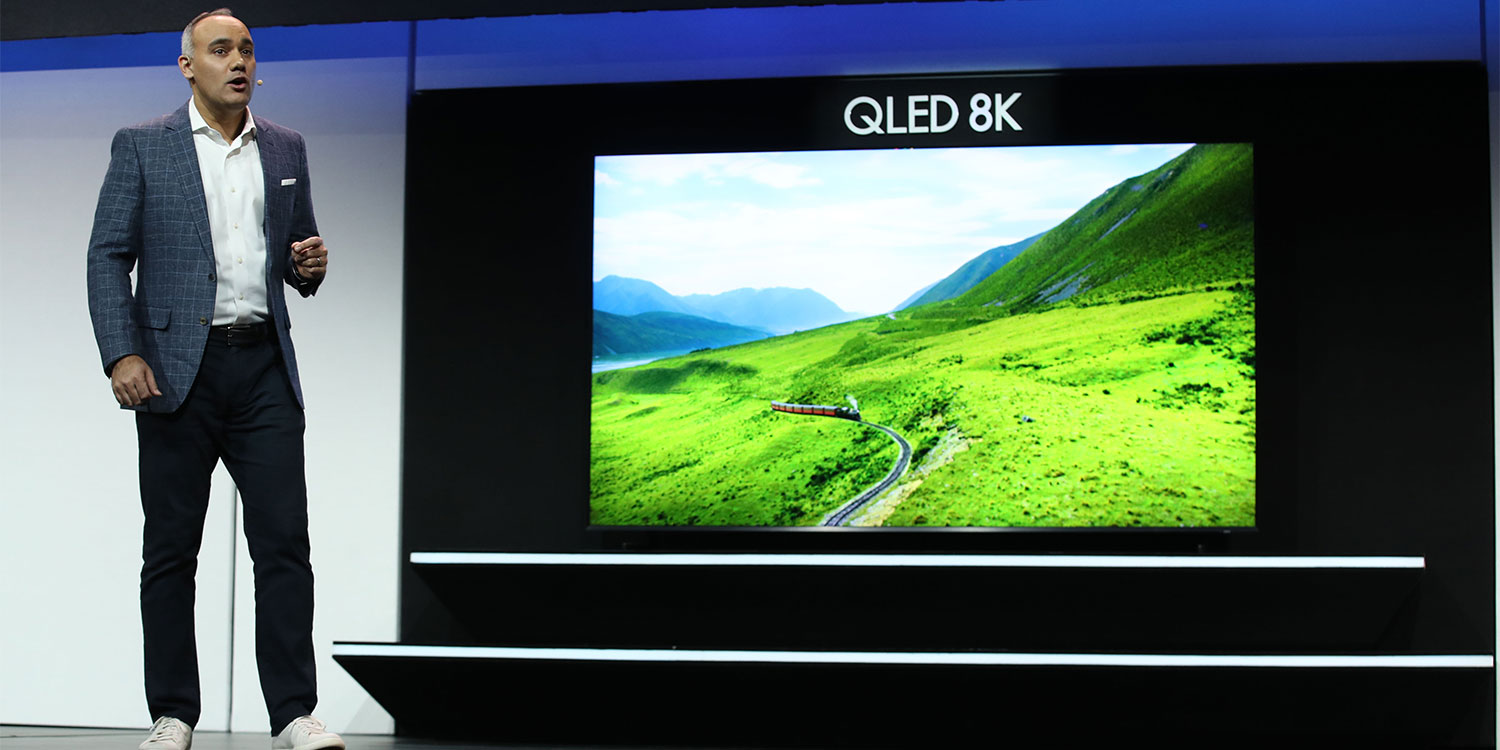 A man in a blazer presents a QLED 8K TV displaying a vibrant green landscape with mountains in the background.
