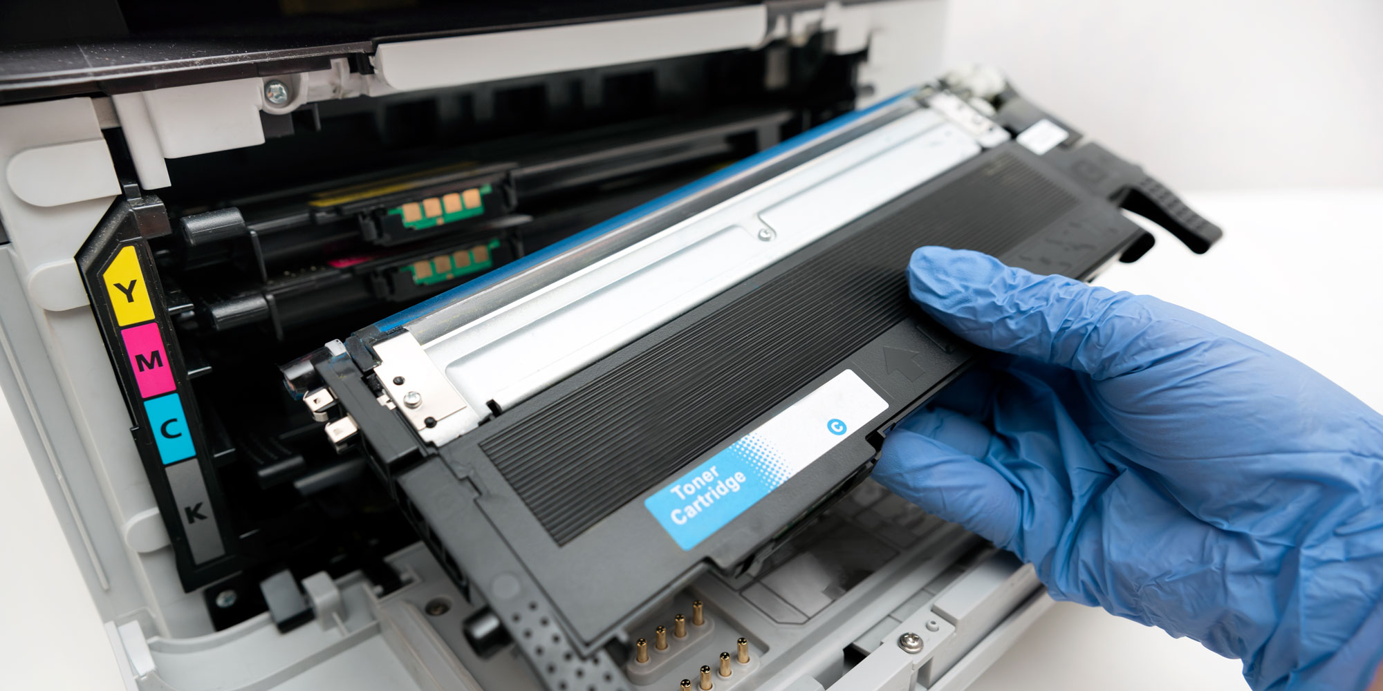 A gloved hand holds a toner cartridge, poised to be inserted into a printer with visible colour labels: Y, M, C, K.
