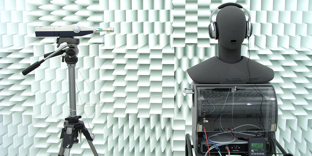 A sound measurement setup featuring a mannequin wearing headphones, a microphone on a tripod, and a textured white wall.