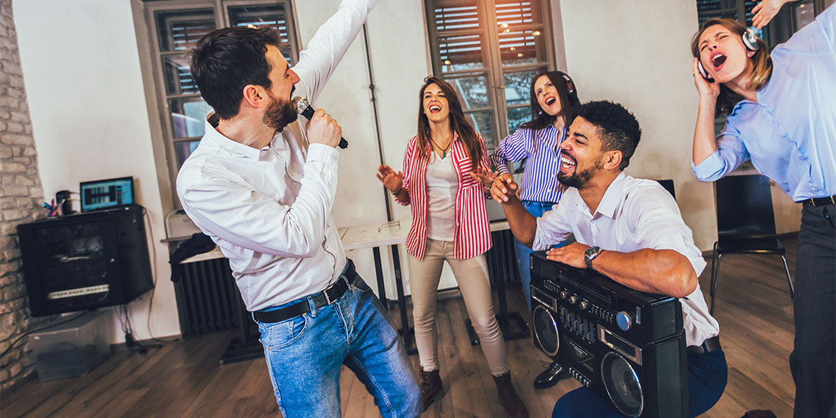 A man sings into a microphone while four friends cheer and dance, some smiling and enjoying the lively atmosphere.