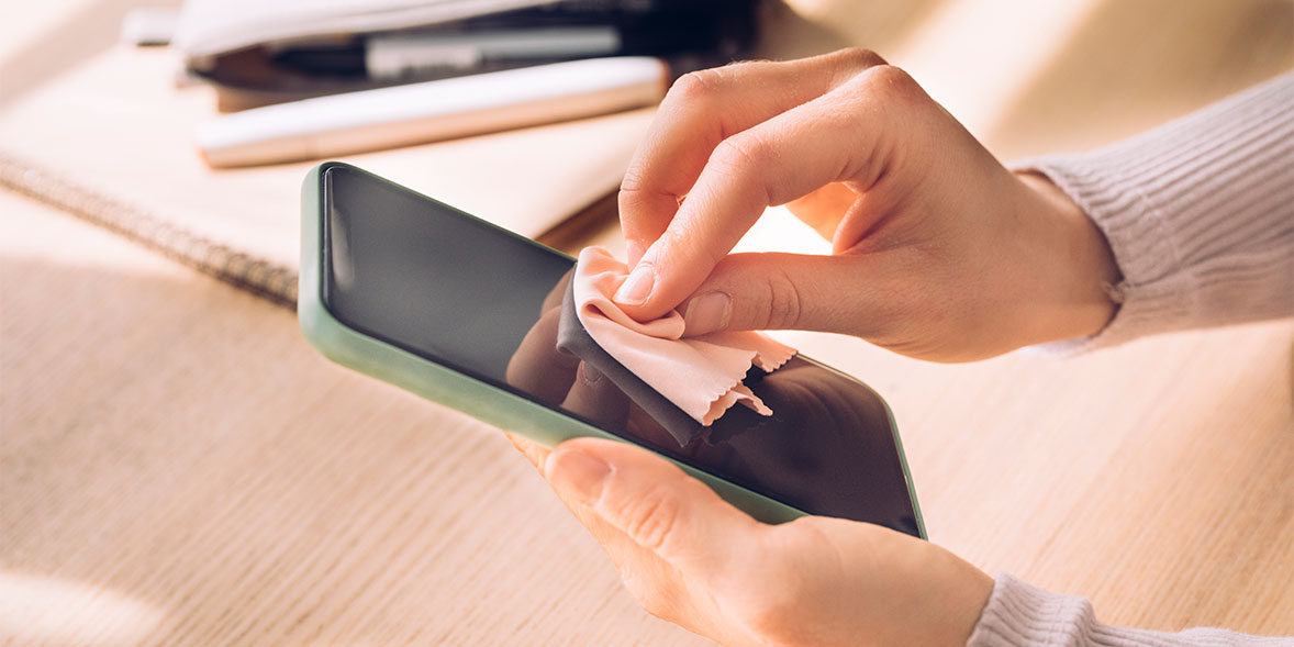 A person wipes the screen of a smartphone with a soft cloth, sitting at a table with notebooks in the background.