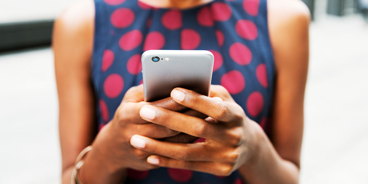 Person in a polka dot dress holds a smartphone, focusing on the screen with both hands, outdoors.