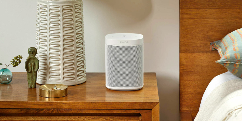 Smart speaker on a wooden table next to a textured lamp and a decorative bowl, with a cozy bedroom background.