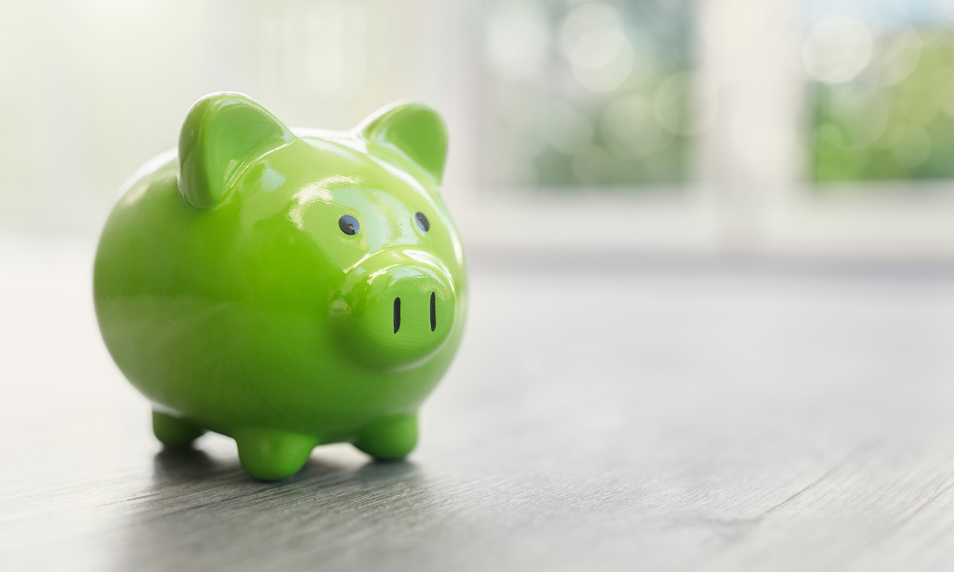 A shiny green piggy bank sits on a wooden floor, with soft natural light coming through a window in the background.