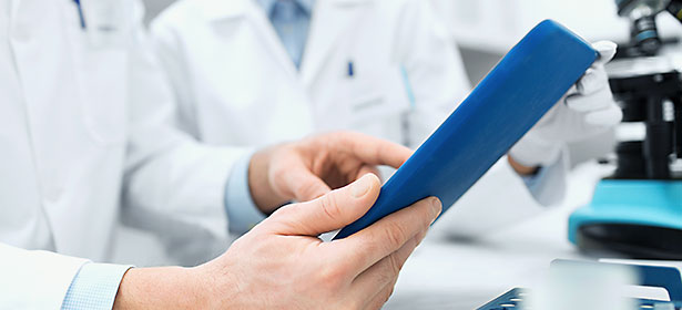 A close-up of hands holding a blue tablet device, with blurred figures in white lab coats in the background.