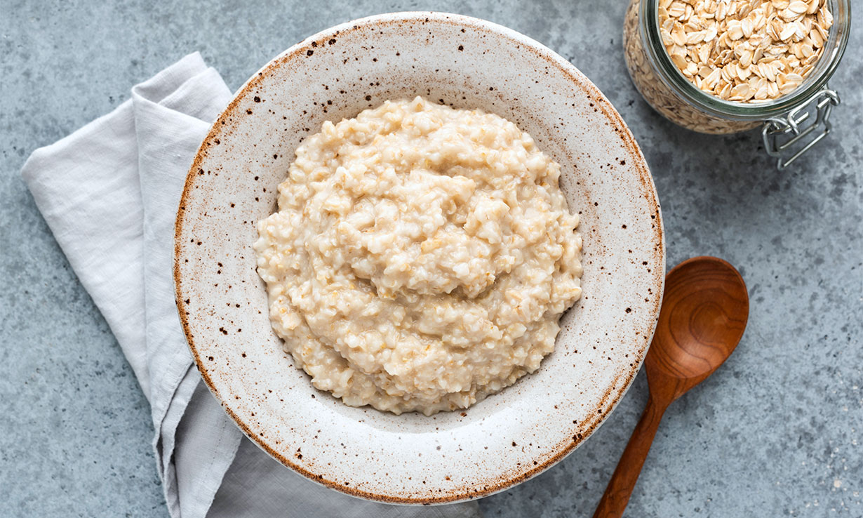 Creamy oatmeal in a speckled bowl, accompanied by a wooden spoon and a jar of oats on a textured countertop.