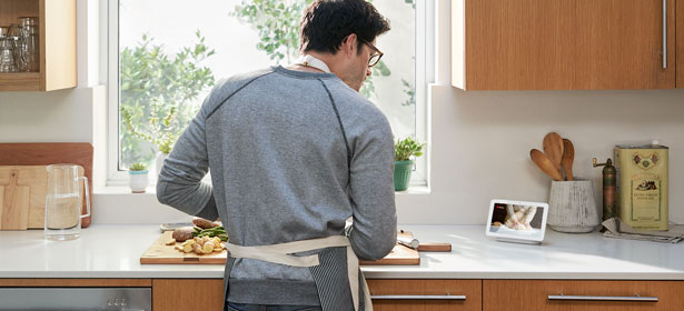 A person in a grey sweater and apron prepares food at a kitchen counter, with a window and tablet displaying a recipe nearby.