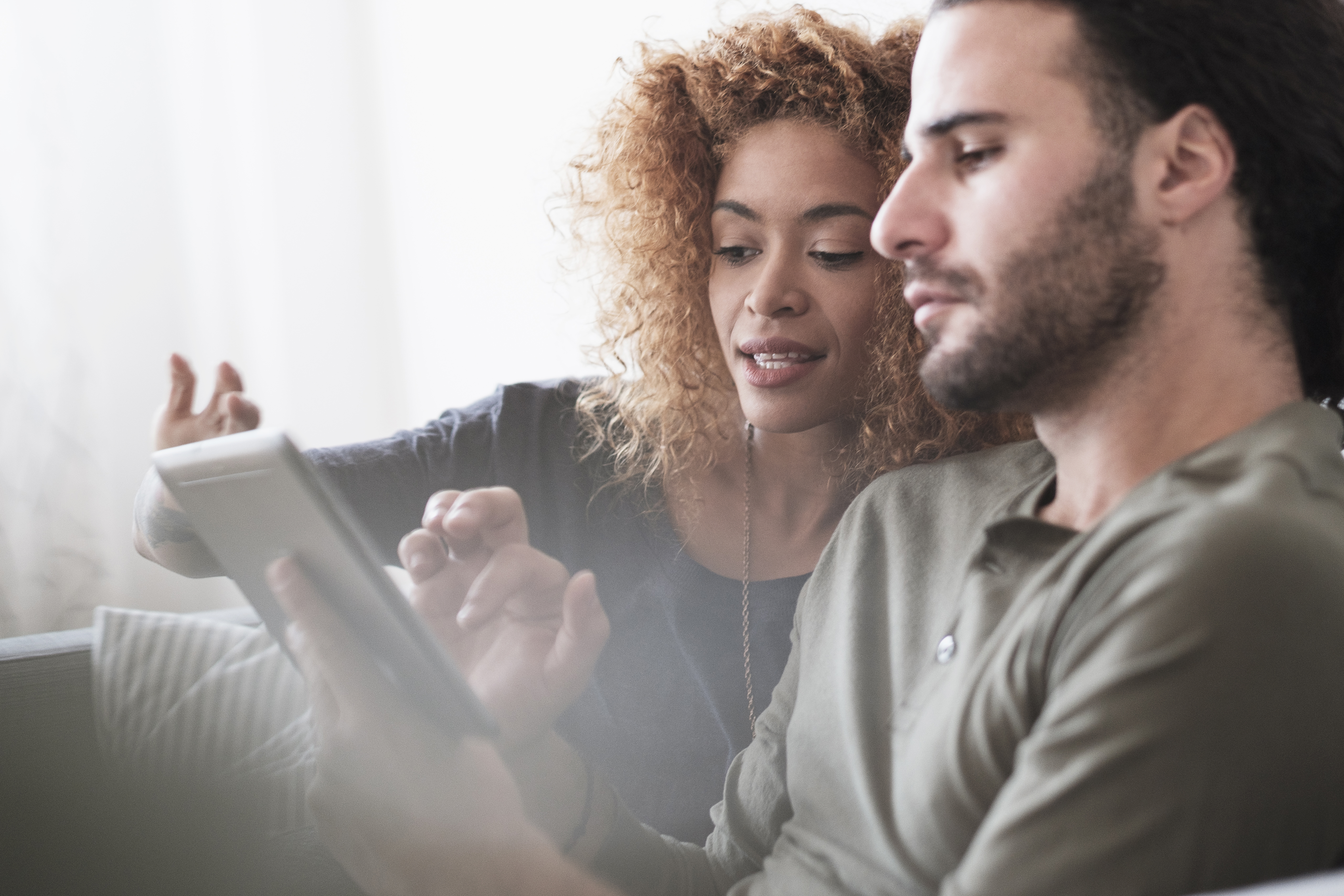 A woman with curly hair enthusiastically points at a tablet while a man focused on the screen sits beside her.