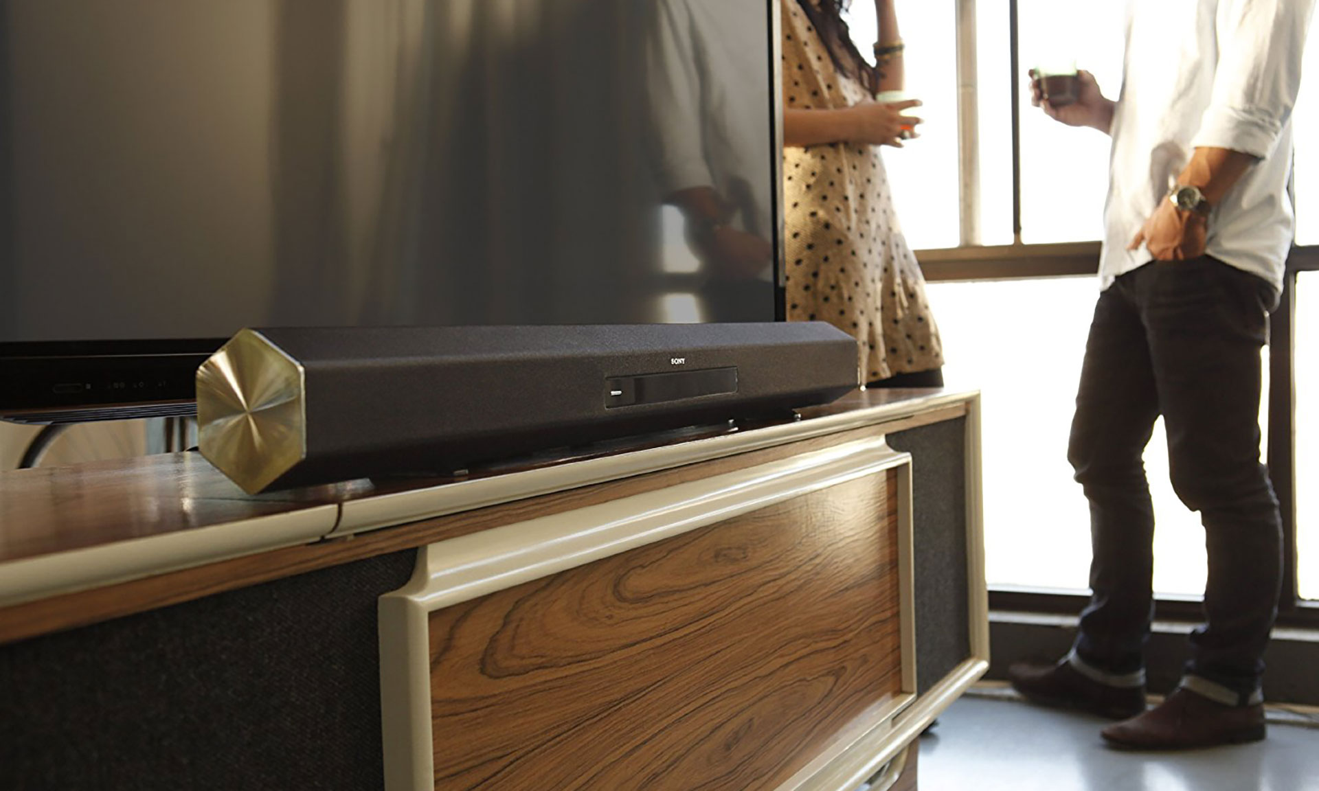 A sleek black soundbar sits on a wooden media console in front of a TV, with two people standing nearby, conversing casually.