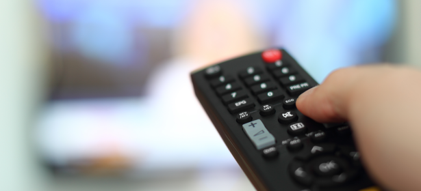 A hand holds a television remote, pointing it towards a blurred TV screen displaying colorful images.
